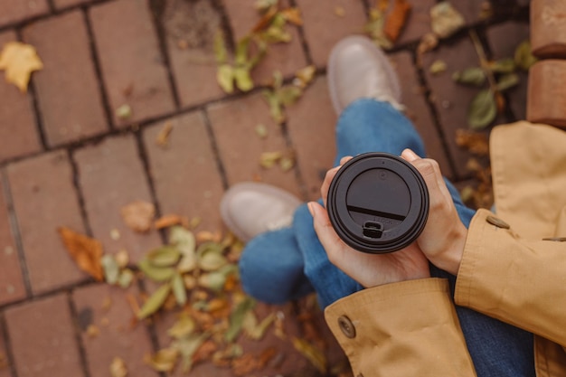 Draufsicht einer Erntefrau, die eine Papptasse mit Kaffee zum Mitnehmen hält und sich die Hände auf einer Bank im Park wärmt