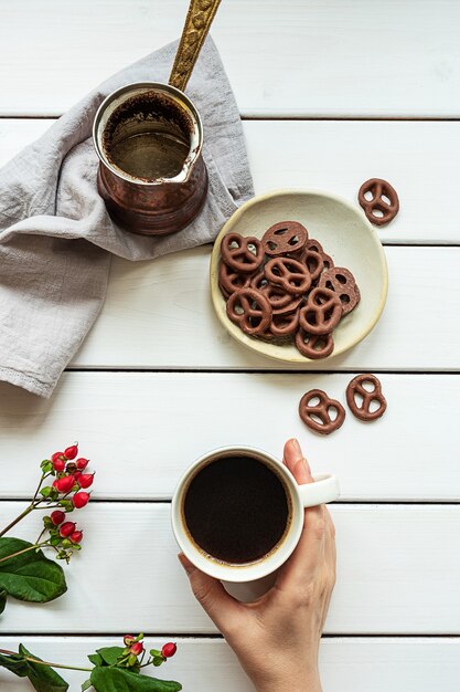 Draufsicht einer hand, die eine tasse kaffee, kaffeekanne und mit schokolade bedeckte brezeln auf einer weißen holzoberfläche hält. zusammensetzung des frühstücks oder der kaffeepause.