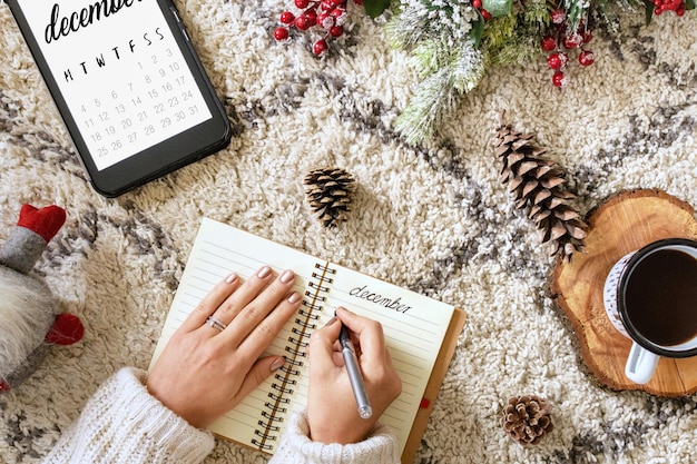 Draufsicht einer kaukasischen Frau, die in einen Planer neben einem Tablet schreibt, das den Kalender zeigt