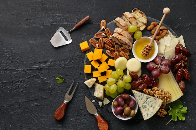 Draufsicht einer leckeren Käseplatte mit Obst, Trauben, Nüssen und Honig auf einer kreisförmigen Küchenplatte auf schwarzem Steinhintergrund, Draufsicht, Kopierraum. Gourmetessen und trinken.