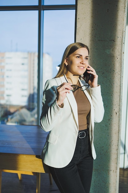 Draufsicht einer positiven Frau in Freizeitkleidung, die in der Nähe eines großen Fensters steht und am Telefon spricht und Informationen während eines Remote-Projekts gibt Moderner Arbeitsbereich für Remote-Arbeit