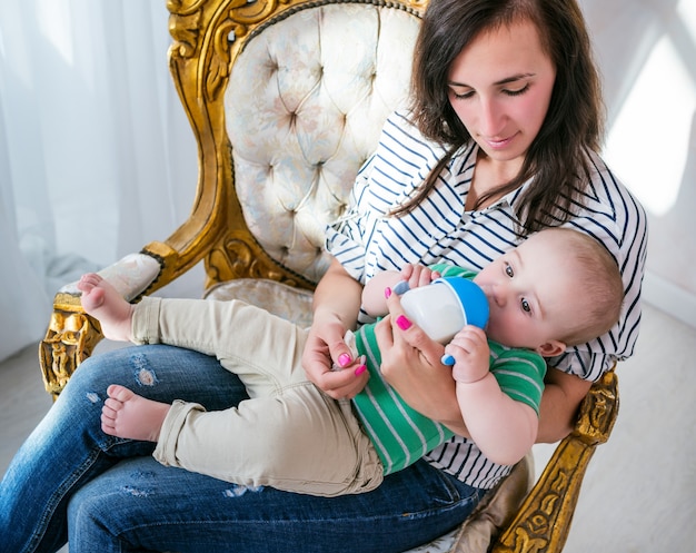 Draufsicht einer positiven jungen Mutter, die ihren kleinen Sohn in ihren Armen hält, während sie auf einem schönen gemütlichen Stuhl im Wohnzimmer sitzt