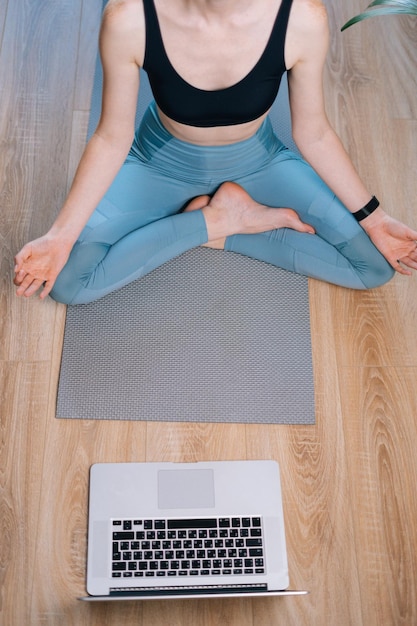 Draufsicht einer unerkennbaren jungen Frau, die zu Hause meditiert und in Lotusposition auf einer Yogamatte in der Nähe eines Laptops sitzt und die Hände auf dem Knie in OM-Position hält.