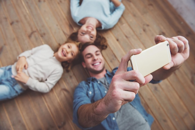 Draufsicht eines hübschen jungen Vaters, der mit seiner schönen Familie ein Selfie macht, während er zu Hause auf Holzboden liegt