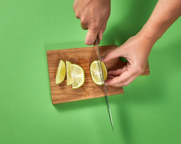 Draufsicht eines Mädchens, das eine Hälfte der organischen reifen Limette mit Scheiben auf einem braunen Brett auf grünem Hintergrund schneidet. Ein scharfes Messer in ihren Händen.