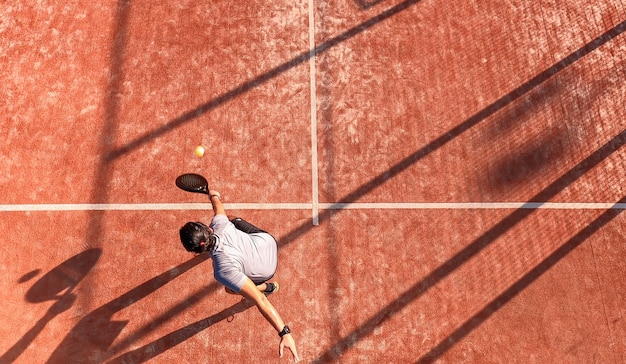 Draufsicht eines Paddle-Tennisspielers, der den Ball mit dem Schläger auf einem Außenplatz schlägt. Er ist in der Nähe der Linie positioniert.