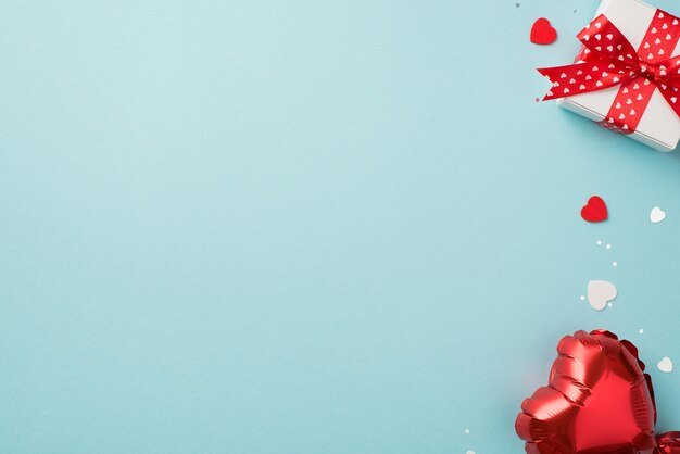 Draufsicht Foto von St. Valentinstag Dekorationen weiße Geschenkbox mit roter Schleife, herzförmigem Ballonkonfetti und Pailletten auf isoliertem pastellblauem Hintergrund mit Copyspace