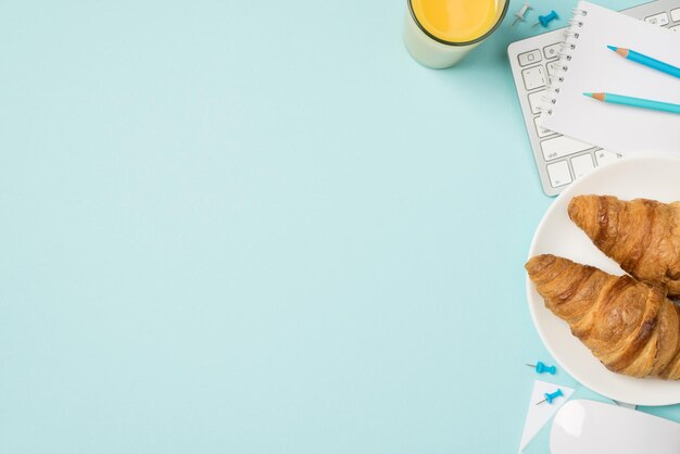 Draufsicht Foto von Tastatur Maus Copybook blaue Bleistifte Stifte Glas Saft und Teller mit Croissants auf isoliertem hellblauem Hintergrund mit Leerzeichen