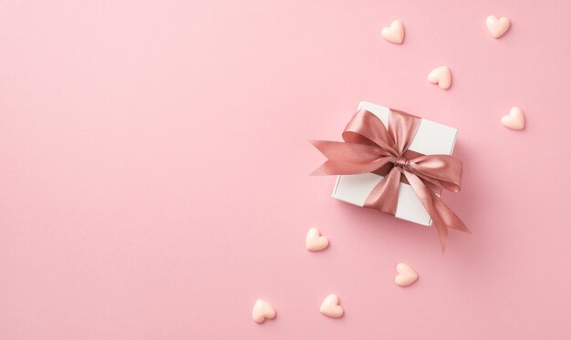 Draufsicht Foto von Valentinstag Dekorationen weiße Geschenkbox mit rosa Seidenschleife und kleinen Herzen auf isoliertem pastellrosa Hintergrund mit Copyspace