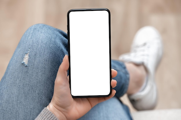 Draufsicht-Mockup-Bild einer Frau, die ein Mobiltelefon mit einem leeren weißen Bildschirm hält Frau in Jeans sitzt mit gekreuzten Beinen und hält ein Mobiltelefon in ihren Händen mit einem leeren weißen Bildschirm