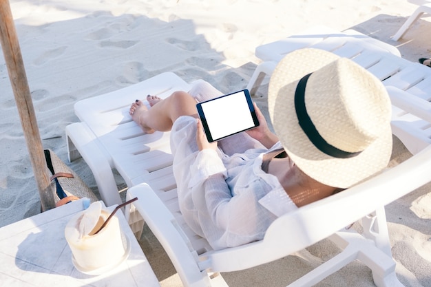 Draufsicht-Modellbild einer Frau, die einen schwarzen Tablet-PC mit leerem Desktop-Bildschirm hält und verwendet, während sie sich auf dem Strandkorb am Strand hinlegt