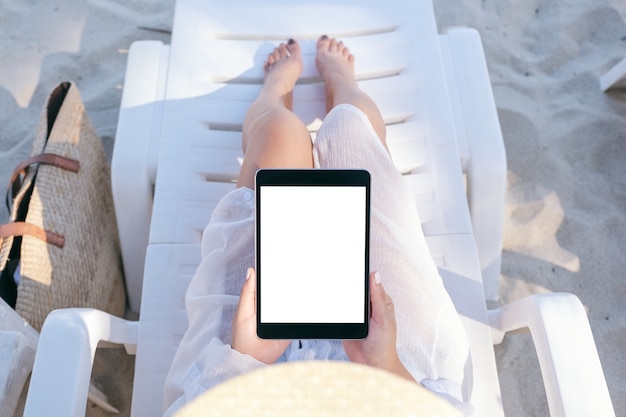 Draufsicht-Modellbild einer Frau, die einen schwarzen Tablet-PC mit leerem Desktop-Bildschirm hält und verwendet, während sie sich auf dem Strandkorb am Strand hinlegt