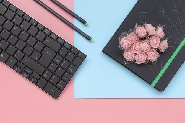 Draufsicht, Notizbuch mit Blumen und Bleistift mit Tastatur auf rosa und blauem Hintergrund.