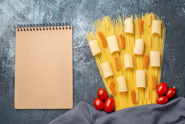 Draufsicht rohe Pasta Rigatoni Spaghetti Spiralen Kirschtomaten lila Schal Notizblock auf grauem Hintergrund