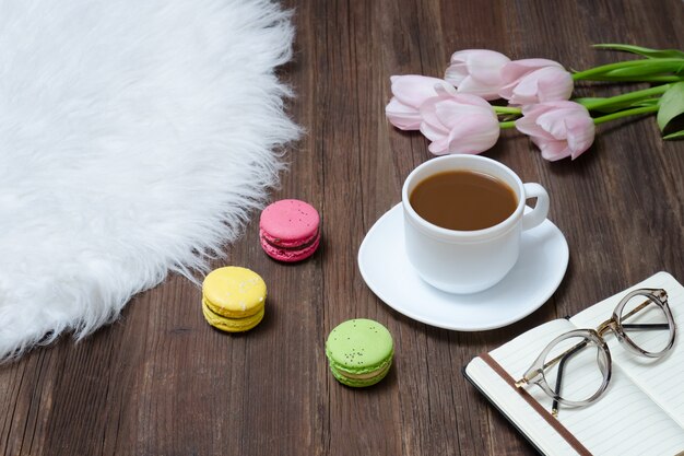 Draufsicht über Tasse Kaffee, macarons, Gläser, rosa Tulpen und Notizbuch auf Holz