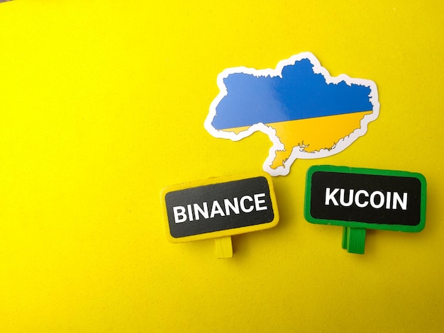 Draufsicht ukrainische Flagge und Holzbrett mit Text BINANCE KUCOIN auf gelbem Hintergrund