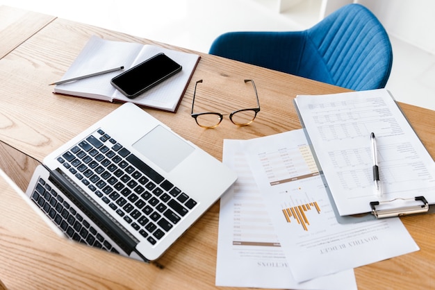 Draufsicht von Büromaterial, das auf Holztisch liegt. Laptop, Zwischenablage, Brille, Bleistift, Notizbuch. Arbeitsplatz mit niemandem