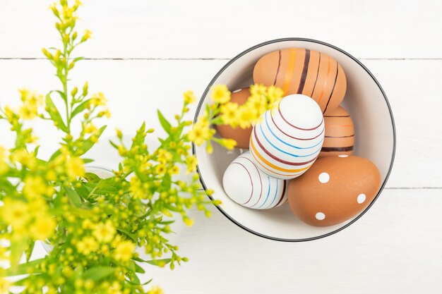 Draufsicht von Ostereiern in einer Schüssel auf hölzernem Hintergrund verziert mit Solidago-Blumen. Gestreifte und gepunktete Ostereier auf einem Tisch.