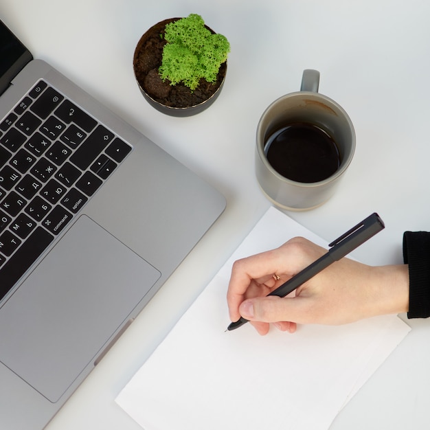 Draufsicht weißer Büroschreibtisch mit leerem Notizbuch, Computer-Laptop-Tastatur, Stift, Pflanze und anderem Büromaterial. Kaffeetasse mit Platz zum Kopieren, flach liegen. Frau Hände mit Telefon.