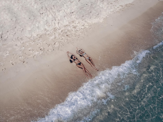 Draufsicht zwei junge Frau in einem Bikini, die am weißen Sandstrand mit türkisfarbenem Meerwasser von Thailand liegt und sich entspannt, Luftbild von der Drohne / Sommerkonzept