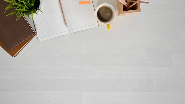 Draufsichtarbeitsplatz mit Buch, Notizbuch, Bleistift und Kaffee auf weißer hölzerner Tabelle.