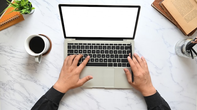 Draufsichtbild der Hände des Geschäftsmannes, die auf Computer-Laptop-Tastatur mit weißem leerem Bildschirm tippen, der auf Marmorbeschaffenheitstabelle setzt, umgeben von Kaffeetasse, Stifthalter, Topfpflanze und Büchern.