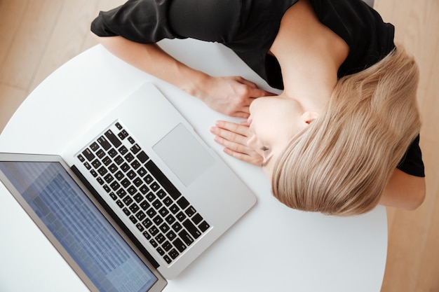 Draufsichtbild der jungen müden Arbeiterin liegt auf dem Tisch im Büro in der Nähe von Laptop-Computern und schläft. Augen geschlossen.