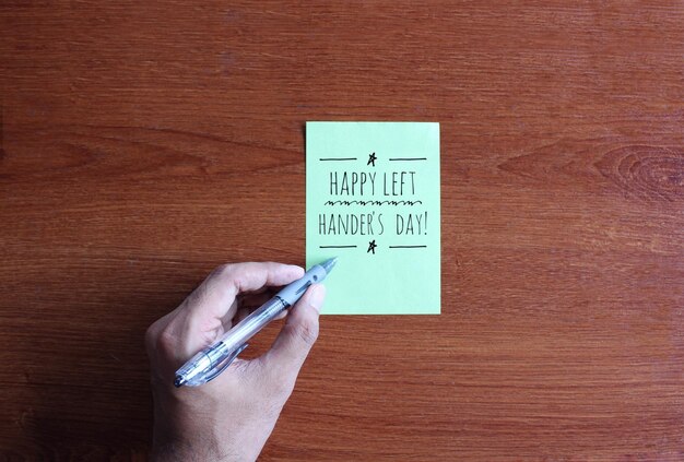 Draufsichtbild der linken Hand schreiben Text Happy Left Hander's Day auf grünem Papier auf Holztisch