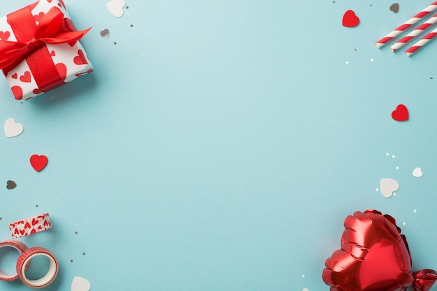 Draufsichtfoto der Geschenkbox mit Valentinstagsdekor in weißem Geschenkpapier mit Herzmuster-Strohhalmen, Klebeband, herzförmigem Ballon, Konfetti, Pailletten auf isoliertem pastellblauem Hintergrund mit Copyspace