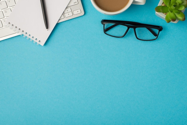 Draufsichtfoto des Desktops mit Tastaturstift auf Notizblock-Brillenpflanze und Tasse Kaffee auf isoliertem blauem Hintergrund mit Leerzeichen