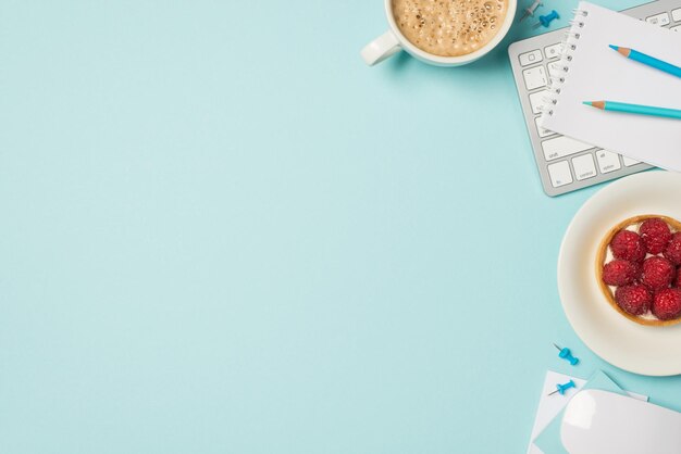 Draufsichtfoto des Tastatur-Maus-Planers mit blauen Stiften, Teller mit Himbeerkuchen und Tasse schaumigem Kaffee auf isoliertem pastellblauem Hintergrund mit leerem Raum