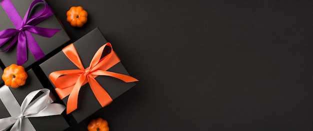 Draufsichtfoto von drei schwarzen Geschenkboxen mit violetten orangefarbenen und weißen Schleifen und kleinen Kürbissen auf isoliertem schwarzem Hintergrund mit Copyspace
