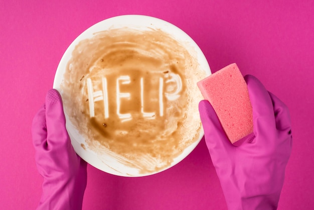 Draufsichtfoto von Händen in rosafarbenen Handschuhen, die weiße schmutzige Schüssel mit Inschrifthilfe und rosa Schwamm auf isoliertem rosa Hintergrund halten