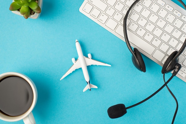 Draufsichtfoto von schwarzen Kopfhörern mit Mikrofon auf weißer Tastaturpflanze, Tasse Kaffee und Flugzeugmodell in der Mitte auf isoliertem pastellblauem Hintergrund