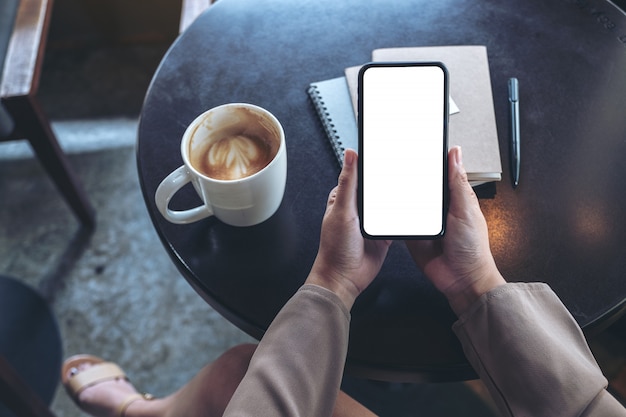 Draufsichtfrau, die schwarzes Handy mit leerem weißen Bildschirm mit Notizbüchern und Kaffeetasse auf Tisch hält