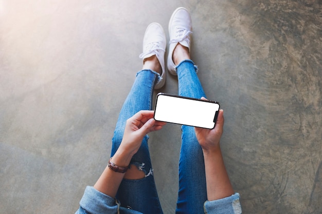 Draufsichtmodell einer Frau, die ein schwarzes Mobiltelefon mit leerem weißem Bildschirm hält, während sie auf dem Boden sitzt
