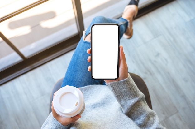Draufsichtmodell einer Frau, die ein schwarzes Mobiltelefon mit leerem weißem Desktop-Bildschirm mit Kaffeetasse hält