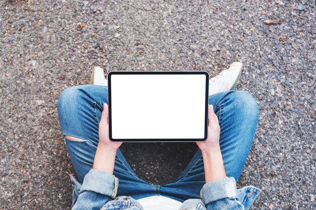 Draufsichtmodell einer Frau, die einen schwarzen Tablet-PC mit leerem Weiß hält, während sie auf der Straße im Freien sitzt