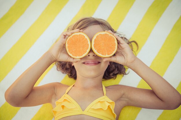 Draufsichtporträt eines Kindes, das Orangenscheiben wie eine Sonnenbrille auf einem gelb gestreiften Handtuch hält