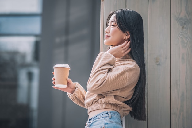 Draußen. Dunkelhaarige junge asiatische Frau mit einer Tasse Kaffee