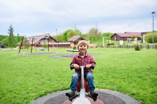 Draußen Porträt des niedlichen lachenden Jungen der Vorschule, der auf einer Schaukel am Spielplatz schwingt