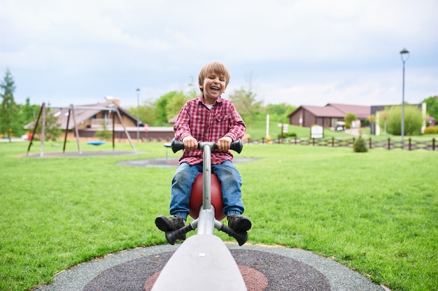 Draußen Porträt des niedlichen lachenden Jungen der Vorschule, der auf einer Schaukel am Spielplatz schwingt