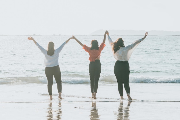 Drei alternative und vielfältige Freunde halten Händchen und haben Spaß am Ufer des Ozeans während eines Sonnenuntergangs. Freunde für immer Freundschaftskonzept. Frauenpower zuversichtlich in die Zukunft