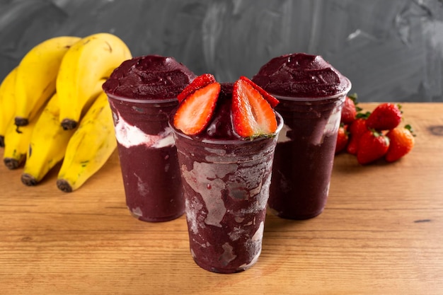 Drei brasilianische Frozen Açai Berry Ice Cream Smoothie in Plastikbecher mit Erdbeeren und Kondensmilch. Auf Holzschreibtisch und grauem Sommerhintergrund mit Früchten. Frontansicht für Speisekarte und Social Media