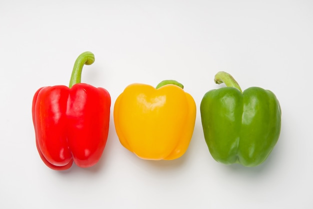 Drei bulgarische Paprika in verschiedenen Farben liegen in einer Reihe auf einer weißen Oberfläche. Sicht von oben.