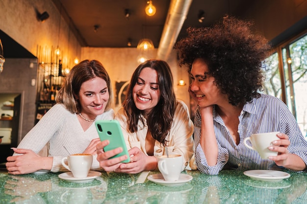 Drei Damen lächeln und haben Spaß mit einem Mobiltelefon, um Textnachrichten im Restaurant oder Café zu senden Eine Gruppe glücklicher junger Frauen, die mit ihrer Handy-App lachen, um Fotos zu teilen