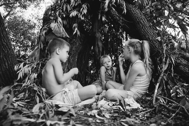 Drei Dorfkinder spielen in einer Hütte, die sie selbst aus Blättern und Zweigen gebaut haben. Holzhaus im Wald. Schwarz-Weiß-Fotografie