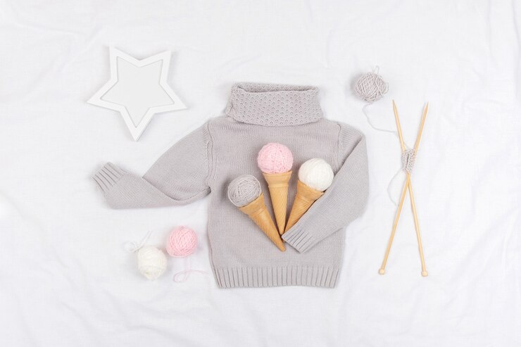 Drei Eiscreme aus Garn und Waffelkegeln auf einem grauen Strickpullover, Holzstricknadeln und Stern auf weißem Hintergrund. Stricken, Hobby und handgemachtes Konzept. Ansicht von oben Flach legen.