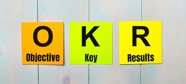 Drei farbige Aufkleber mit dem Text OKR Objective Key Results auf hellblauem Holzhintergrund