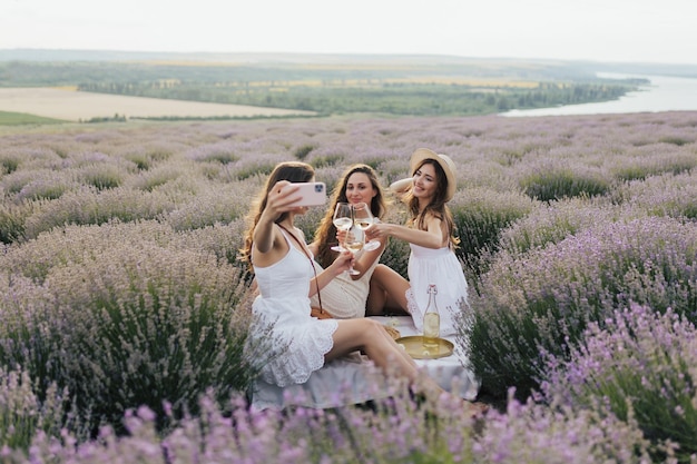 Drei Frauen in weißen Kleidern sitzen in einem Lavendelfeld, eine davon hält ein Glas Wein in der Hand.